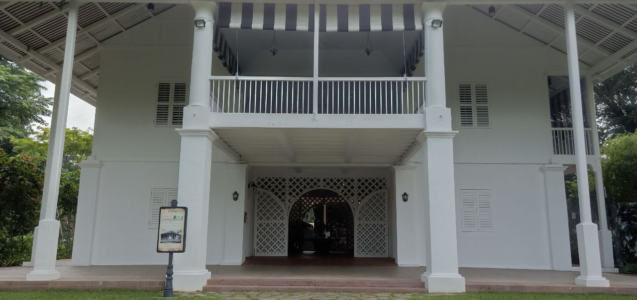 a white colonial style building with white pillars and a white awning over the entrance. the building has a balcony on the second story and a porch on the ground floor. the windows are shuttered and the door is arched. there is greenery surrounding the building.
