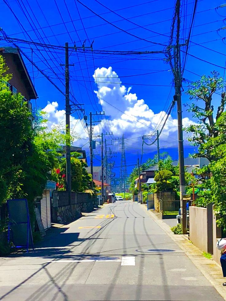 a view down a residential street with houses on either side, trees lining the road, and a bright blue sky with puffy white clouds overhead. power lines and utility poles are visible above the street.
