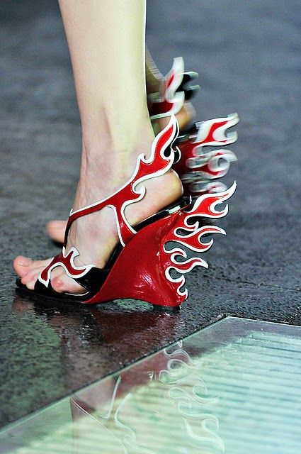 a closeup shot of a person's foot wearing a red and white sandal with a flame design, on a glossy surface. the sandal has a wedge heel and is adorned with stylized flame shapes. the leg is positioned vertically, and the shoe is the primary focus, highlighting its unique design.
