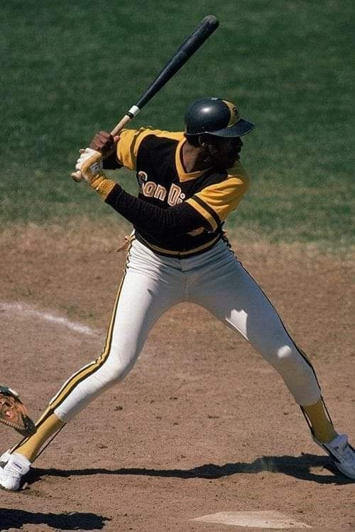 a san diego padres baseball player in a black and yellow uniform is caught midswing, bat connecting with the ball. the image is taken from an eyelevel perspective on a sunny day. the player's focus and determination are evident.
