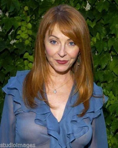 a closeup portrait of a woman with long, red hair, wearing a blue top with a ruffle collar and sheer sleeves, smiling for the camera. she has a necklace on and the background is blurry.  the photo was taken in a studio.