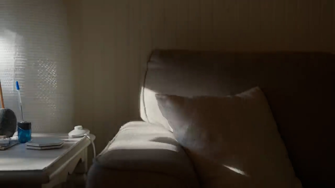 a sunlit living room featuring a beige couch with a white pillow, a white side table with a book and a pen, and a window with white curtains.