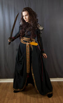a fulllength shot of a woman dressed in a dark, elegant fantasy warrior costume. she is holding a katana and standing on a wooden floor against a dark gray background. her long brown hair is loose, and she is wearing a long black robe with gold detailing and a yellow sash. the costume appears intricately detailed, suggesting a historical or fantasy setting. her expression is serious and focused, enhancing the powerful and fierce image she projects. the overall mood of the image is dark and dramatic.