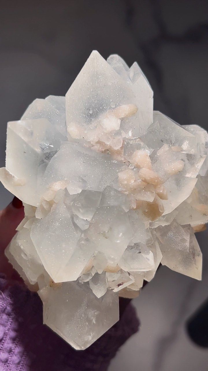 a closeup photo of a cluster of clear, white crystals. the crystals are translucent and shiny, and they have a rough, natural texture. the crystals are pointed and clustered together in a beautiful, random formation. there are some hints of pink color in the center of the cluster. the background is blurred out of focus.  the crystals are in a hand holding it.