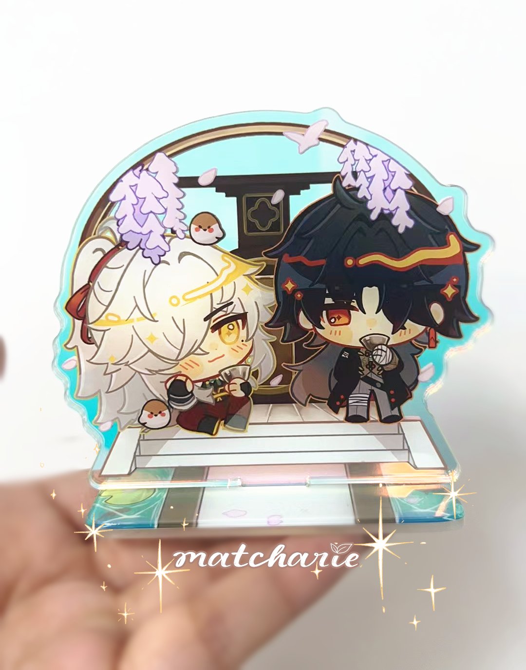 a close up of a clear acrylic stand with a  chibi anime figurine on it. the figurine has  white hair, and a  red, yellow, and white outfit. the  figure is  sitting on a bench with another figurine, who has black hair and a black and white outfit. the stand is translucent and features the characters sitting on a bench under a flowered trellis. there are small birds on the bench. it is in the palm of someone's hand, which is slightly blurred. the stand has a signature in the bottom that says "matcharie".