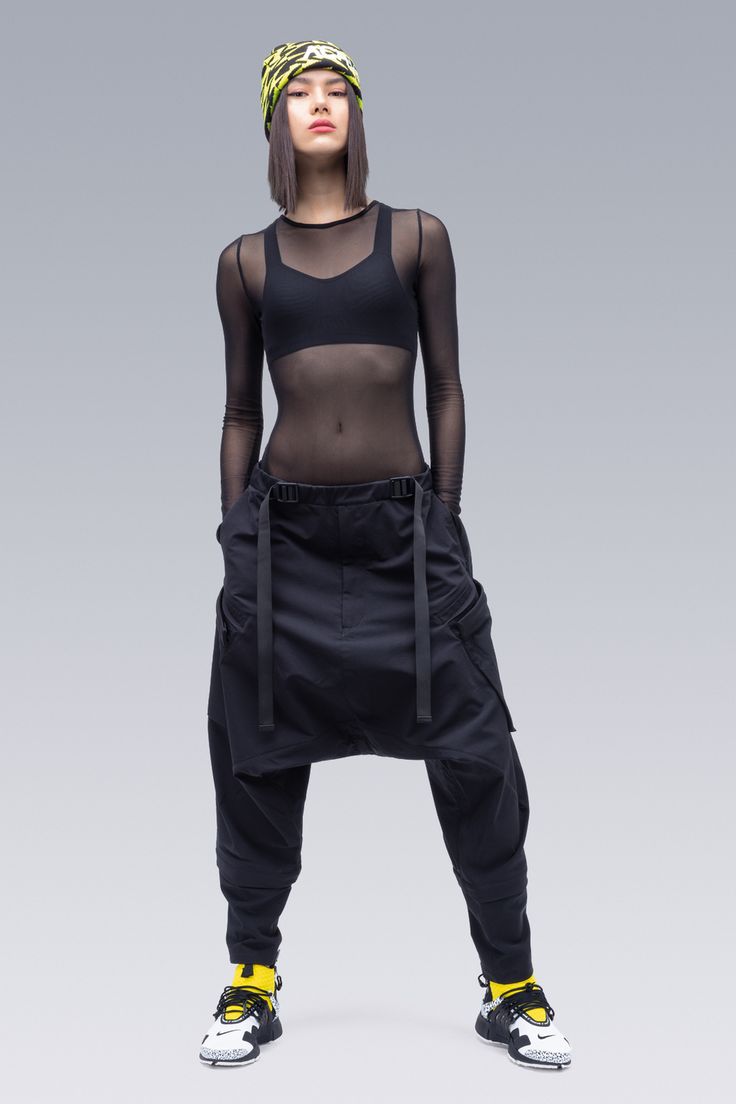 a fulllength studio shot of a woman standing and facing the camera, showcasing a fashionforward athletic outfit. she wears a black mesh top over a black sports bra, paired with black cargo pants that feature distinctive front straps. bright yellow socks add a pop of color and are visible above her white and black athletic shoes. a yellow and black patterned headband completes the look. the background is a solid, light grey.