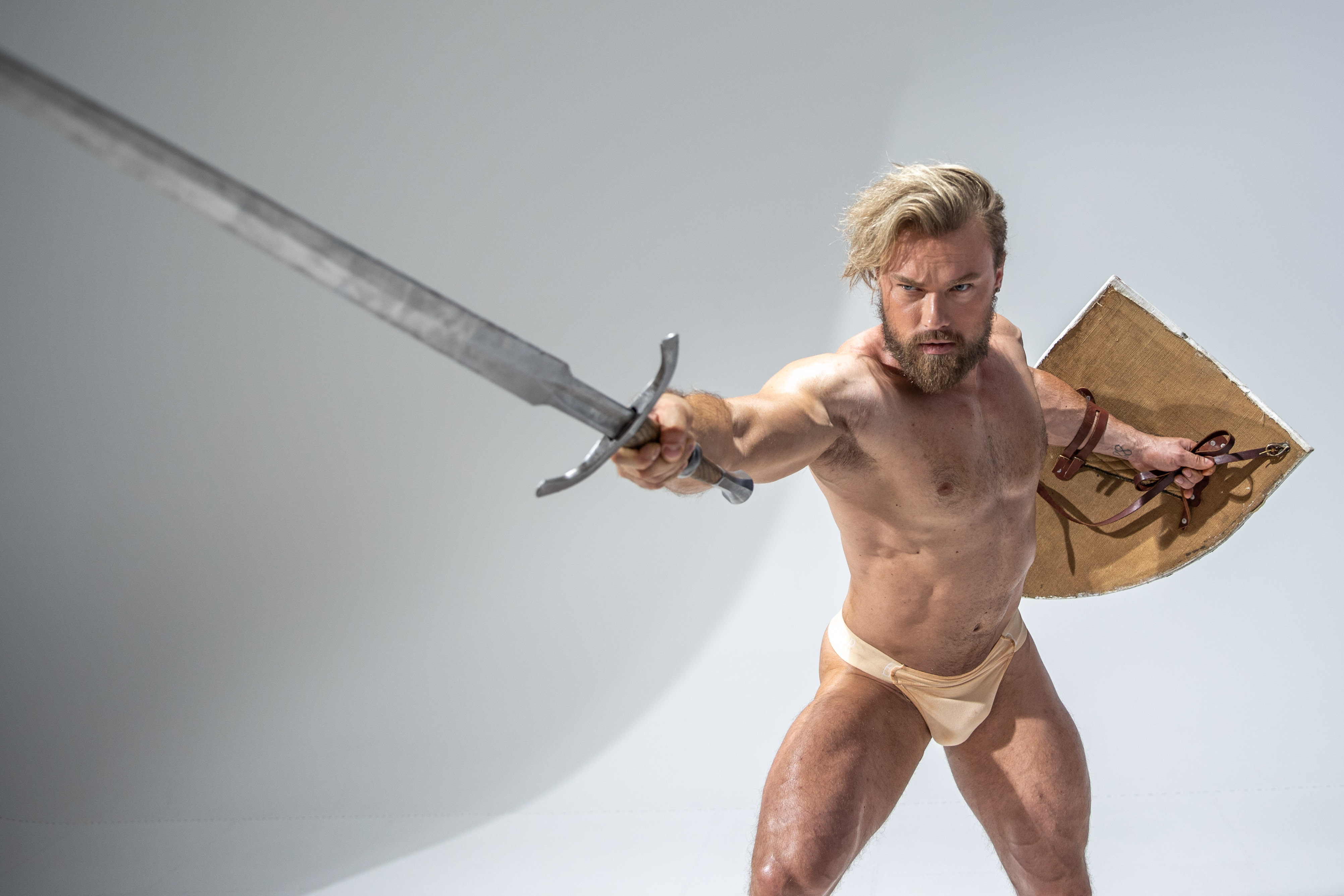 a muscular man with blonde hair and beard  in a fighting stance while wielding a sword and shield.  his physique is on full display as he wears only a loin cloth,  leather arm band and brown leather strapped shield. he appears to be preparing to attack.