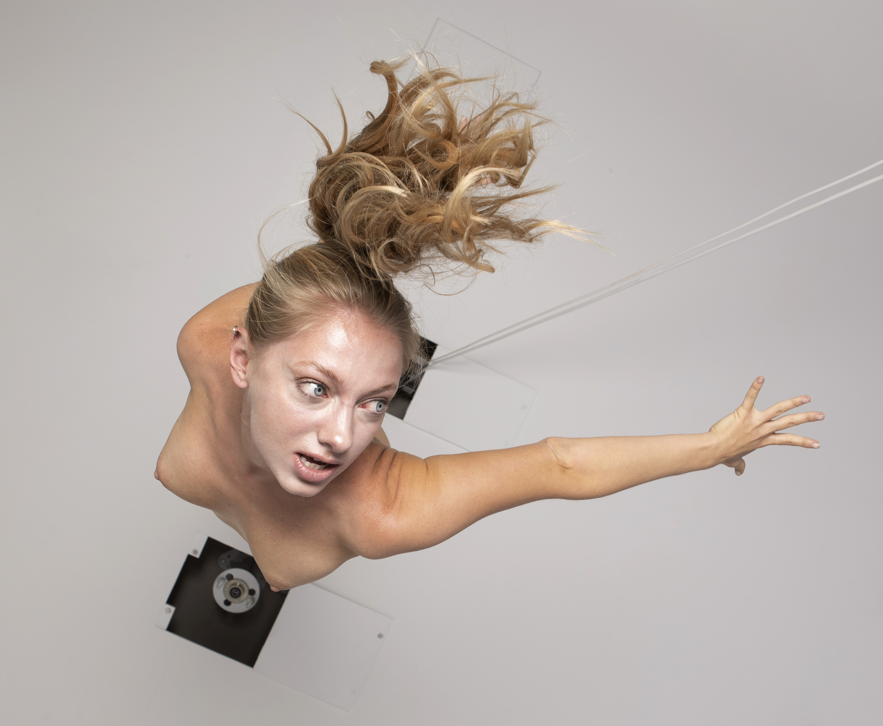 a lowangle shot of a fairskinned woman with blonde hair in a high bun, looking away with a surprised expression. she has bare shoulders and is positioned as if emerging from a hole in a white ceiling. mechanical structures are visible around the opening, along with a few wires. she is reaching out with one arm. the overall composition is artistic and creates an unusual perspective.