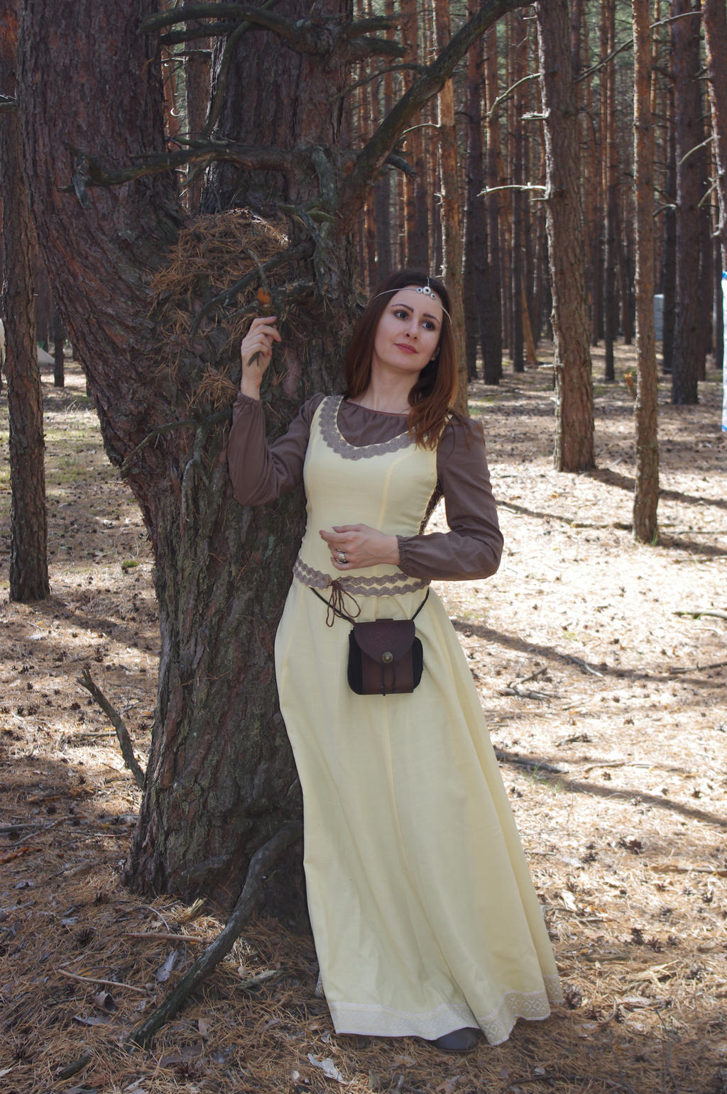 a woman in a long, pale yellow medievalstyle dress stands in a pine forest. she wears a brown overdress and a headband. she carries a small brown purse.  the sunlight filters through the trees. the scene has a fantasy or fairytale atmosphere.
