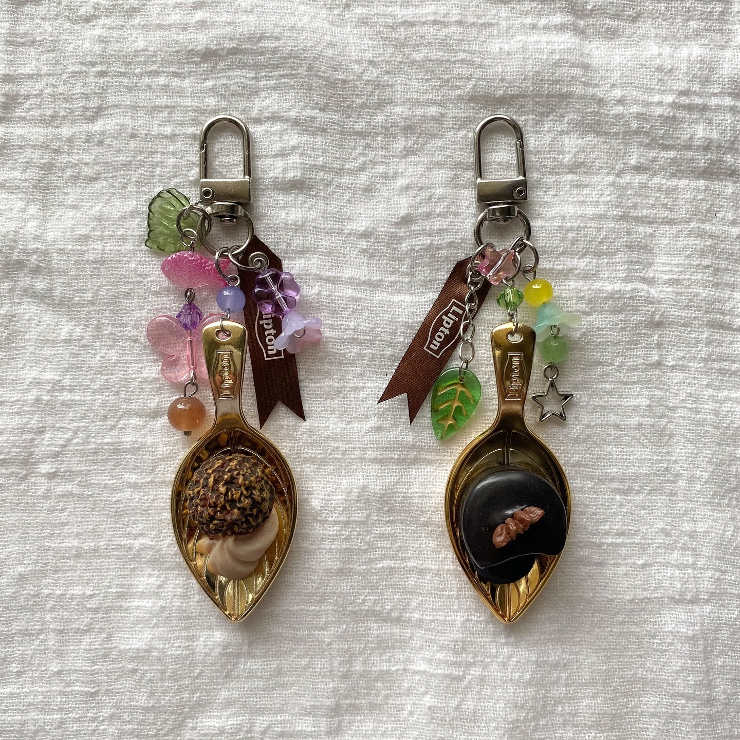 two keychains are displayed on a white textured surface. each keychain consists of a golden spoon holding a piece of chocolate or candy. various beads, charms (including a star and a leaf), and a small ribbon labeled "lipton" adorn each keychain.