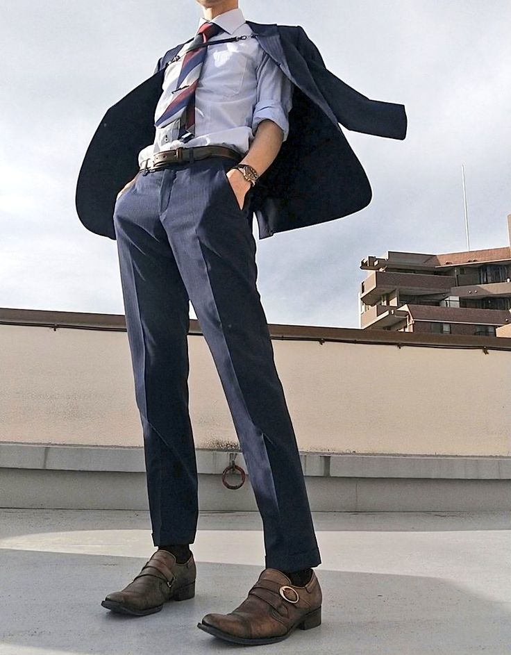 a man is standing on a rooftop, wearing a sharp navy blue suit. he is wearing a white dress shirt, and a red, white, and blue striped tie. he is also wearing brown leather shoes with buckles. he looks confident and stylish.