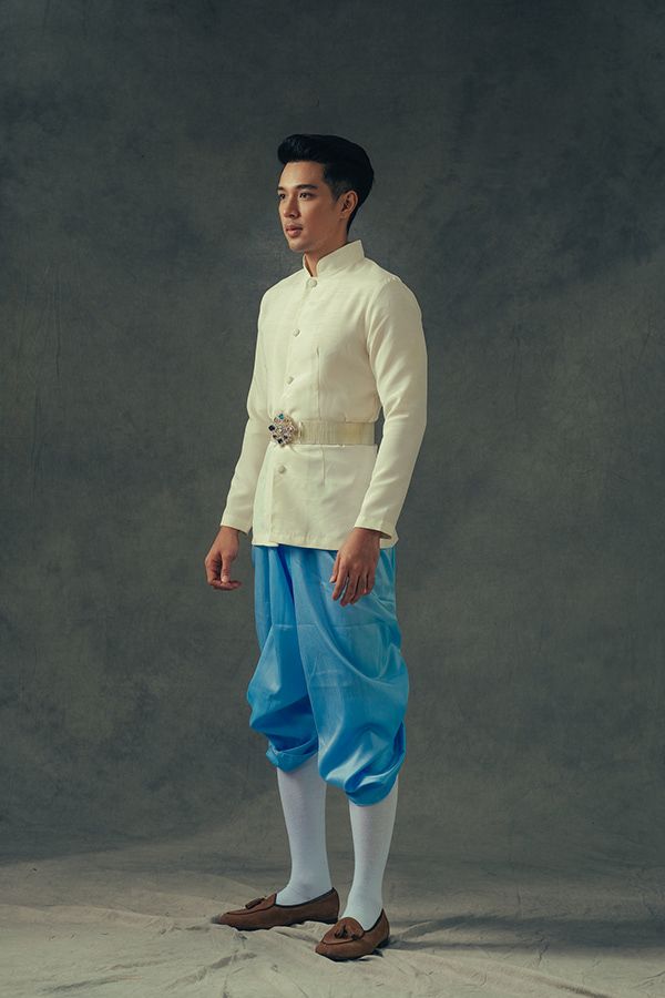 a man wearing traditional thai clothing, consisting of a white shirt, blue pants, white socks, and brown shoes, stands in a studio with a grey background. he is looking off to the left side of the frame.