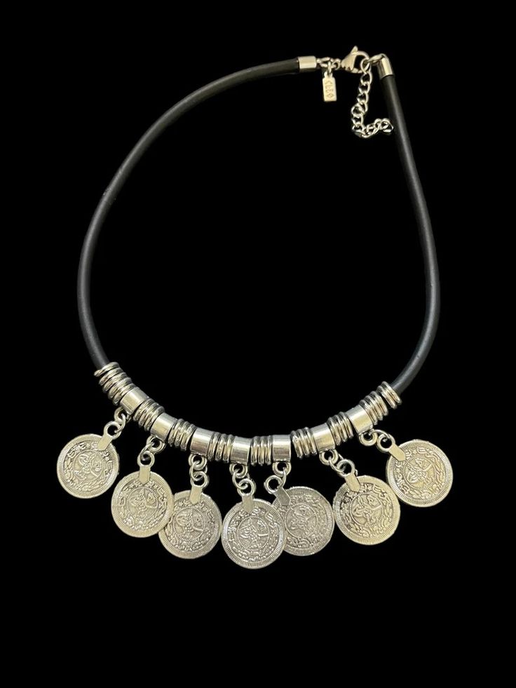 a silver coin necklace with a black leather cord. the necklace has 7 silver coin pendants that are ornate and have arabic script on them. the necklace is isolated on a black background.