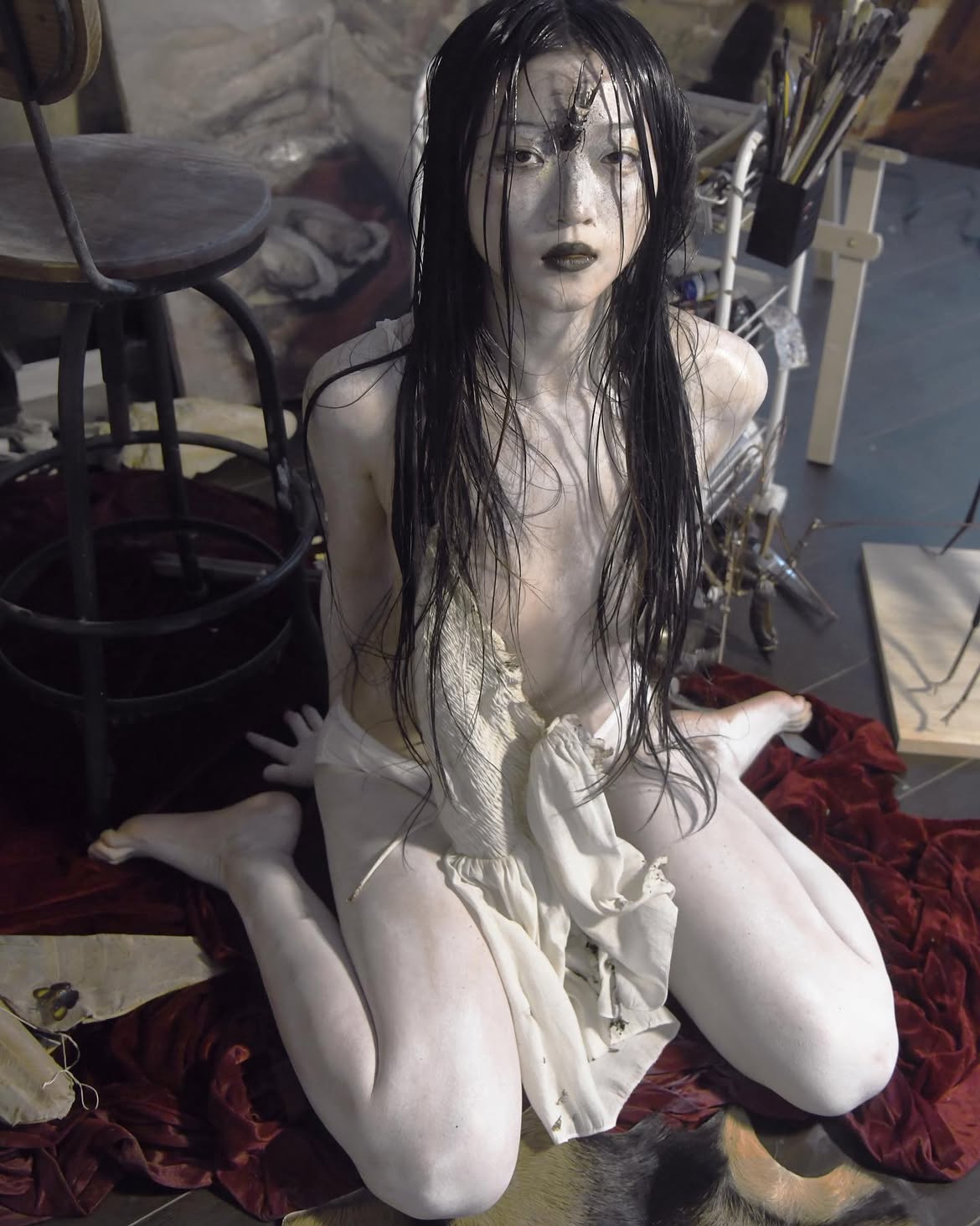a paleskinned woman with black lipstick and wet black hair kneels on a red fabriccovered floor in a studio setting. her skin appears to be painted, and she has a hornlike object affixed to her forehead. she is partially covered by white cloth. the setting includes a stool, artwork, and painting supplies, suggesting a surreal art installation or performance art piece. the overall impression is bizarre, unconventional, and otherworldly.