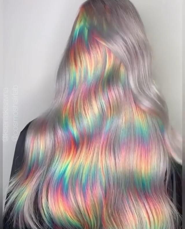 a person with long, silver hair that is iridescent, changing colors in the light. the colors are a blend of pinks, blues, greens, and yellows. the hair is styled in loose waves.  the person is wearing a black shirt.