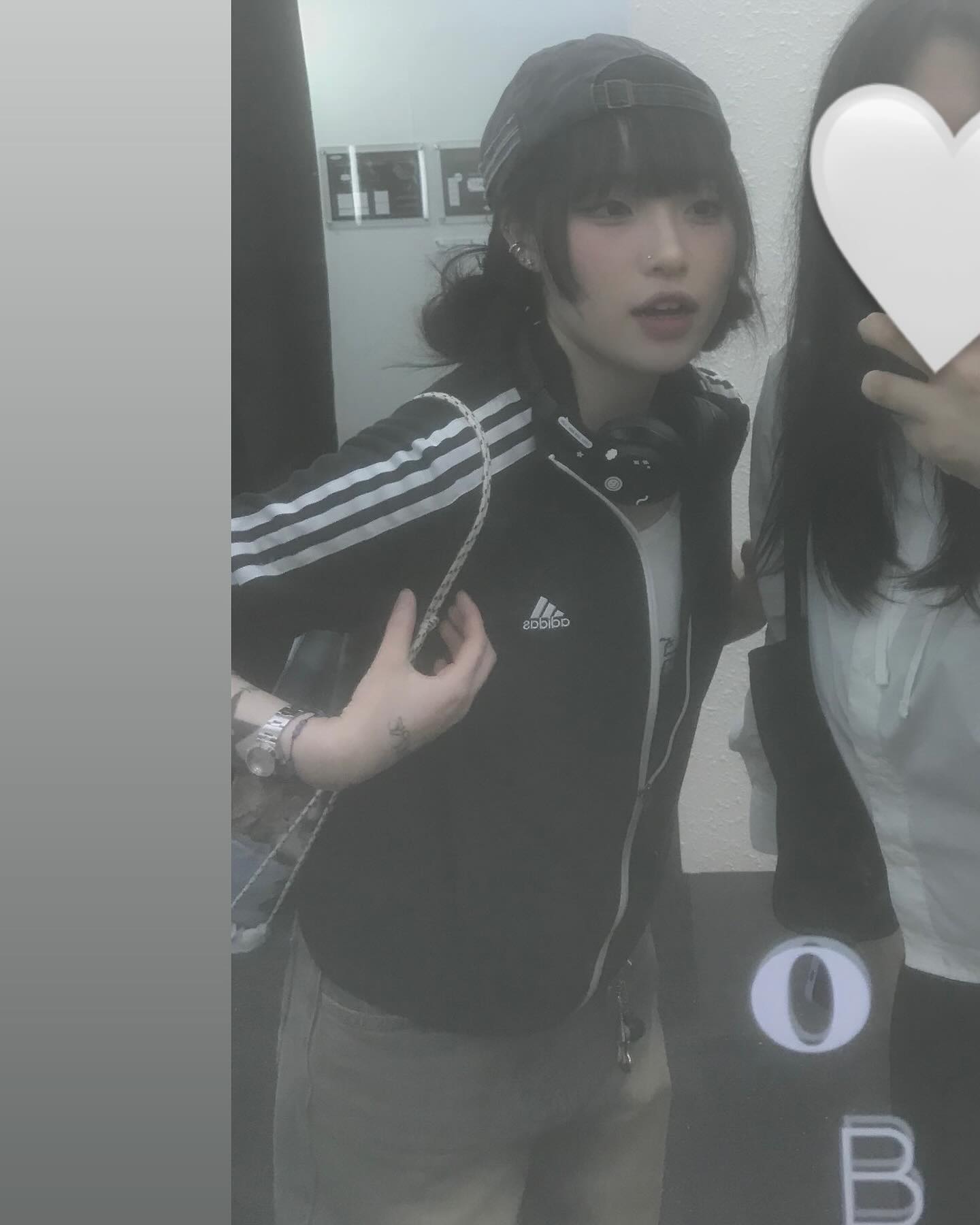 the image shows two women taking a mirror selfie. the woman in the foreground is wearing a black adidas track jacket, a black cap, and light grey pants. her hair is black and styled in a short, edgy cut. she has several tattoos visible on her arms. the second woman is partially obscured and wearing a white hoodie. the background suggests they are indoors, possibly in a hallway or backstage area. the lighting is artificial and slightly dim, creating a grainy, almost nostalgic feel.