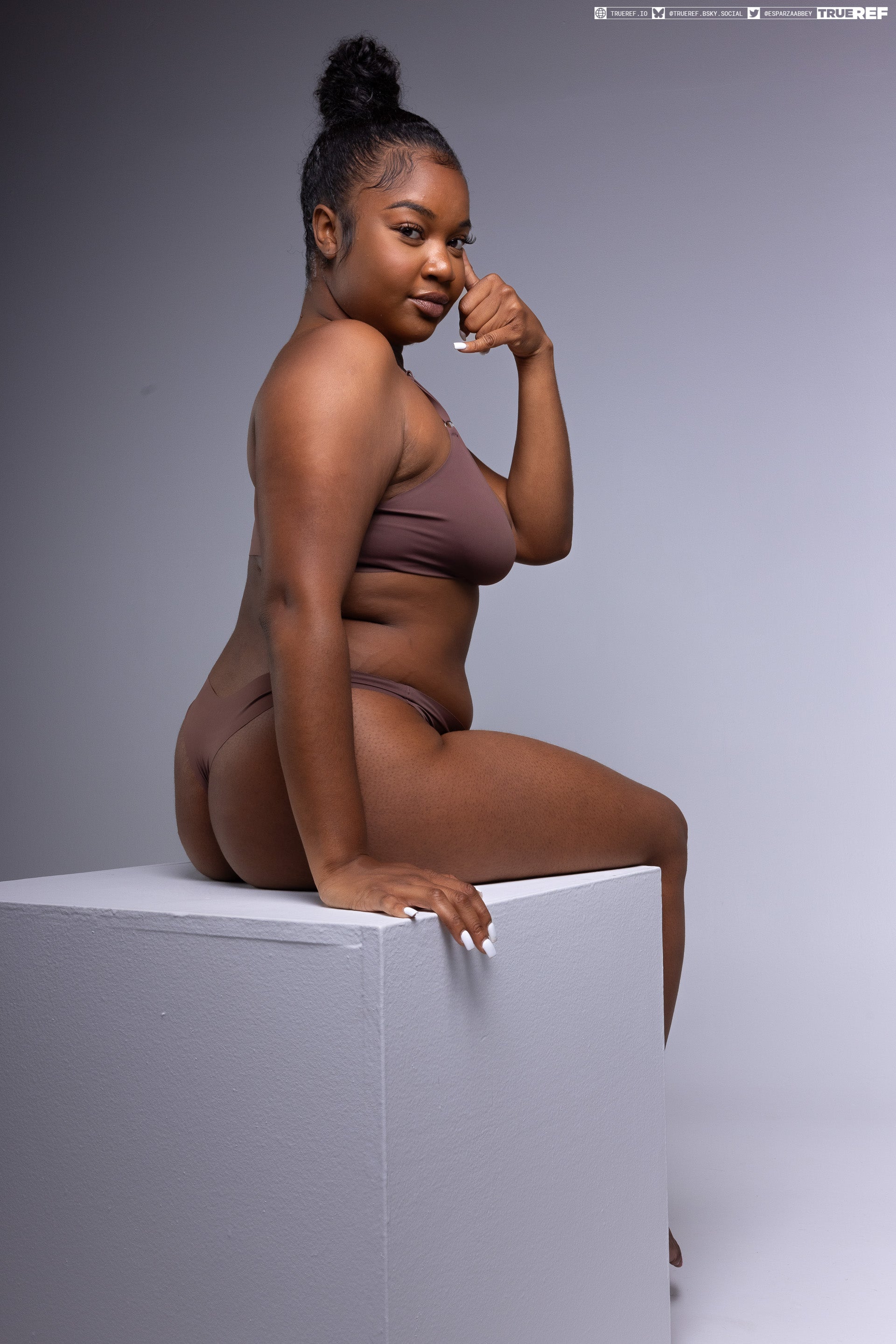 a confident woman is sitting on a white cube in a studio setting. she is wearing brown underwear and has her dark hair styled in a bun. she is looking directly at the camera with a confident expression and has one hand on her face. the background is a plain light gray, which puts the focus entirely on her. the scene is welllit, with a studio shot quality.