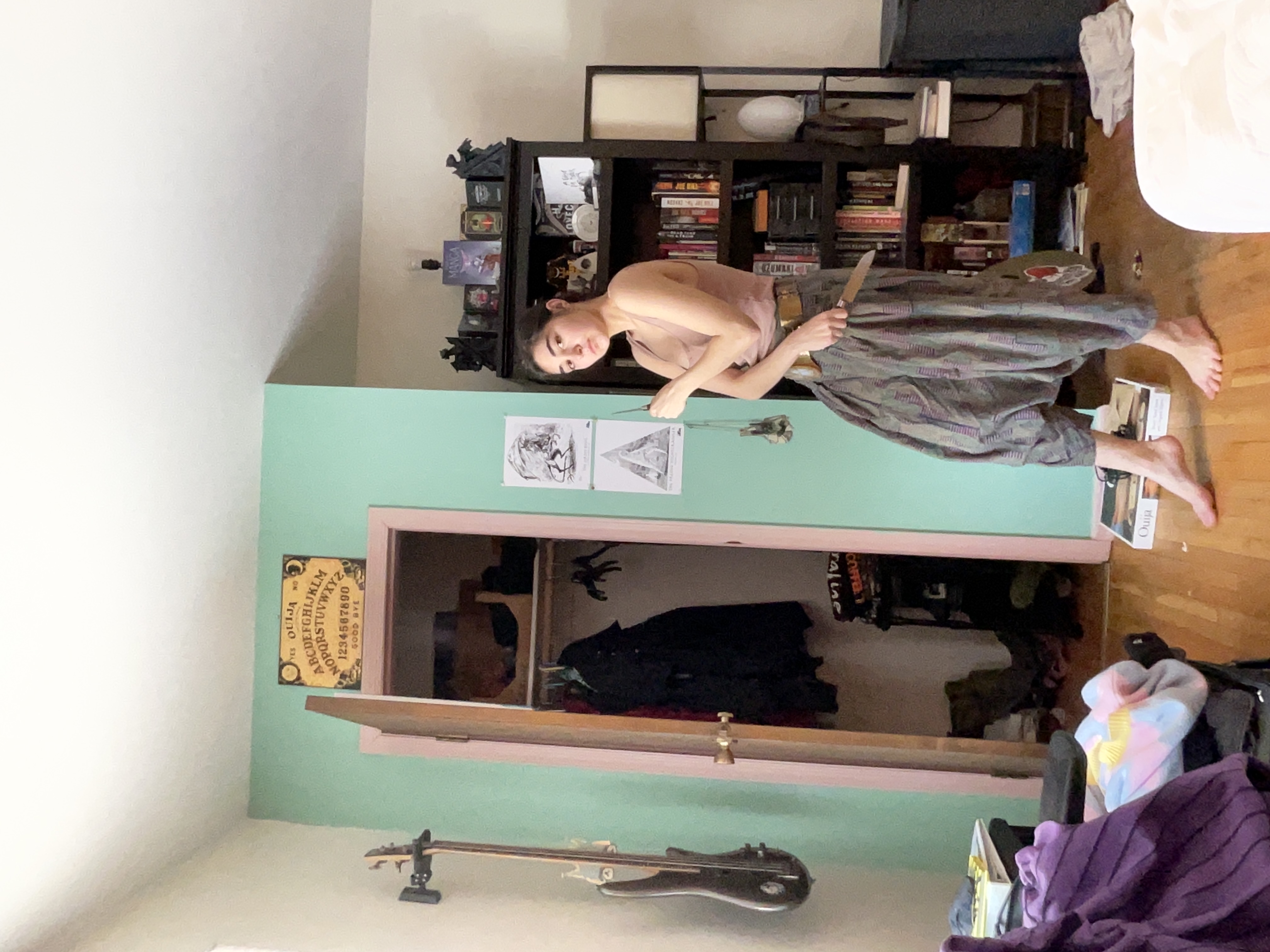 a young woman stands in her bedroom, holding a knife. she's wearing harem pants and a tank top. the room is eclectically decorated, with a bookshelf filled with books, a guitar leaning against the wall, and a ouija board hanging on the wall. a closet is visible in the background, along with a wheelchair partially in view. the overall style of the room suggests an interest in occult or mystical themes.