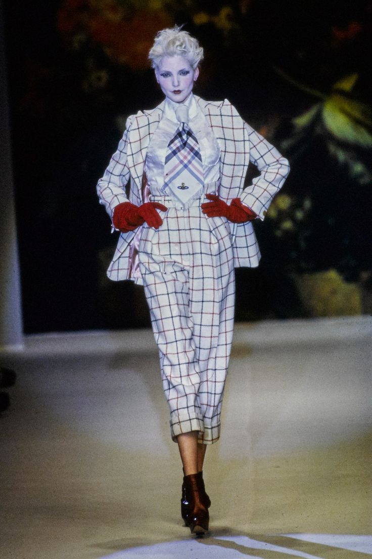 a female model walks down a runway in a white and black checkered suit. she has a red tie and gloves on, and is wearing brown high heels. her hair is blonde and she is wearing makeup.