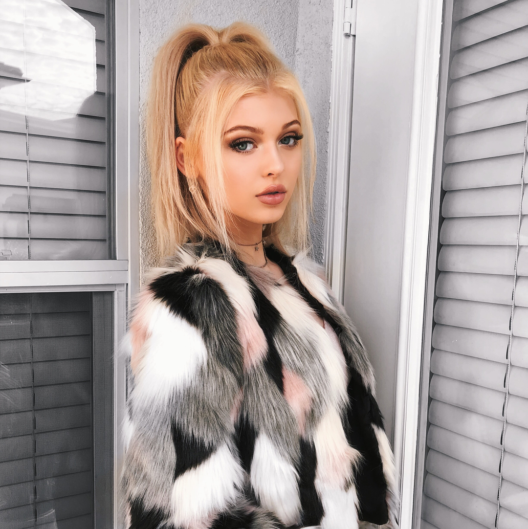 a young woman with blonde hair styled in a high ponytail is standing in front of a window. she is wearing a fur coat with a pattern of black, white, pink, and gray. she has makeup on and is wearing jewelry, including a choker. the window has blinds.
