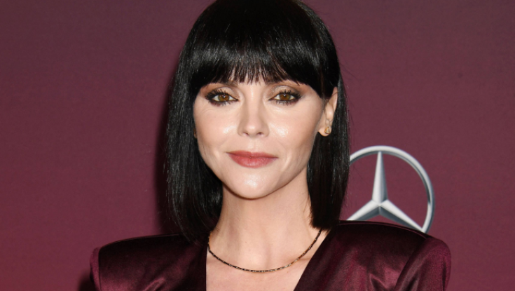 closeup portrait of actress rose mcgowan. she is wearing a maroon dress and a simple necklace. her hair is styled in a black bob with bangs. she's looking directly at the camera with a neutral expression. the background is a deep burgundy color with a mercedesbenz logo visible in the upper right corner. the image appears to be from a red carpet event or photoshoot.