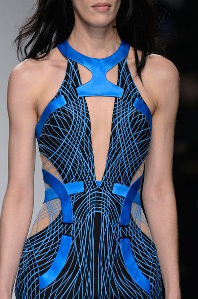 a woman model is wearing a long, blue and black dress on a runway. the dress has a geometric pattern and cutout design. the model has long dark hair and her makeup is dramatic with dark eyes and bold lips. she is posing for the camera and the dress is fitted to her figure. the lighting is dramatic and the background is blurred.