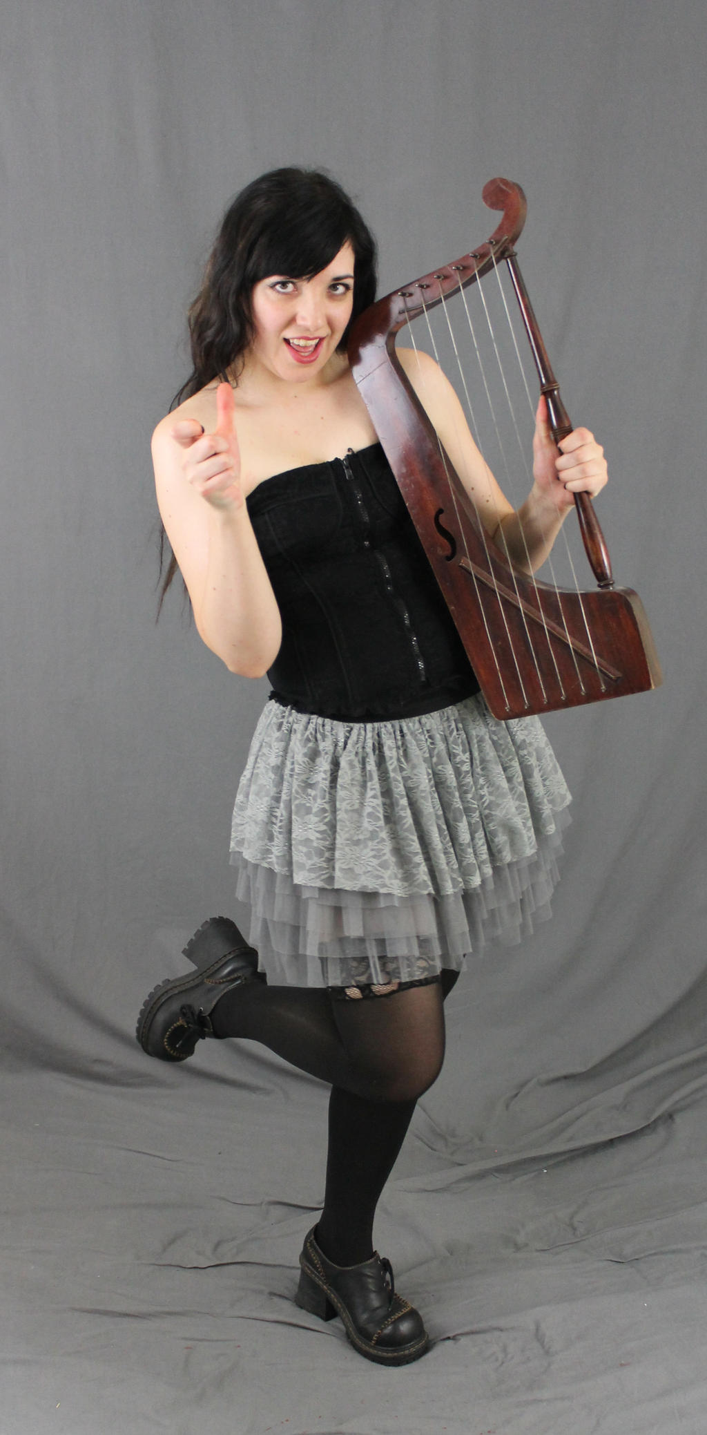 a young woman in a black corset and grey skirt is playing a harp. she is smiling and giving a thumbs up. she is wearing black stockings and black shoes. the background is grey.