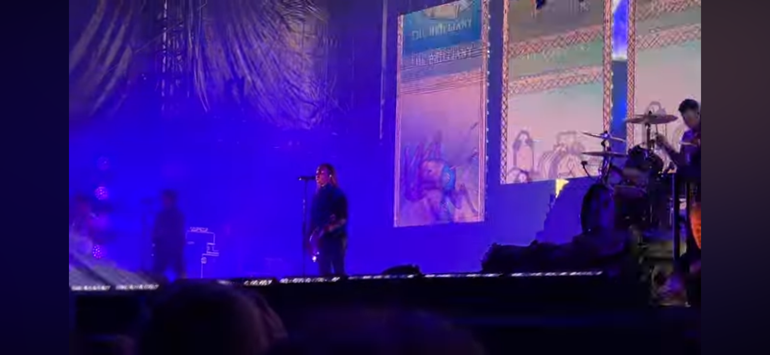 a live concert with a band performing on stage. the lead singer is center stage with a microphone and guitar, the drummer is on the right, and a guitarist is on the left. the stage is lit with purple and blue lights, and there is a large screen behind the band with abstract designs. the audience is in the foreground, with their backs to the camera.