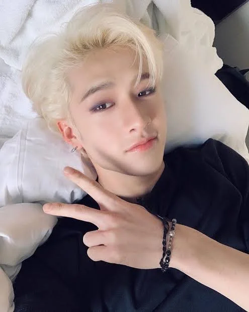 a young asian male with blond hair is lying on a bed with a white pillow. he is wearing a black shirt and a bracelet on his right wrist. he is smiling and giving a peace sign. the image is a closeup portrait of the man.