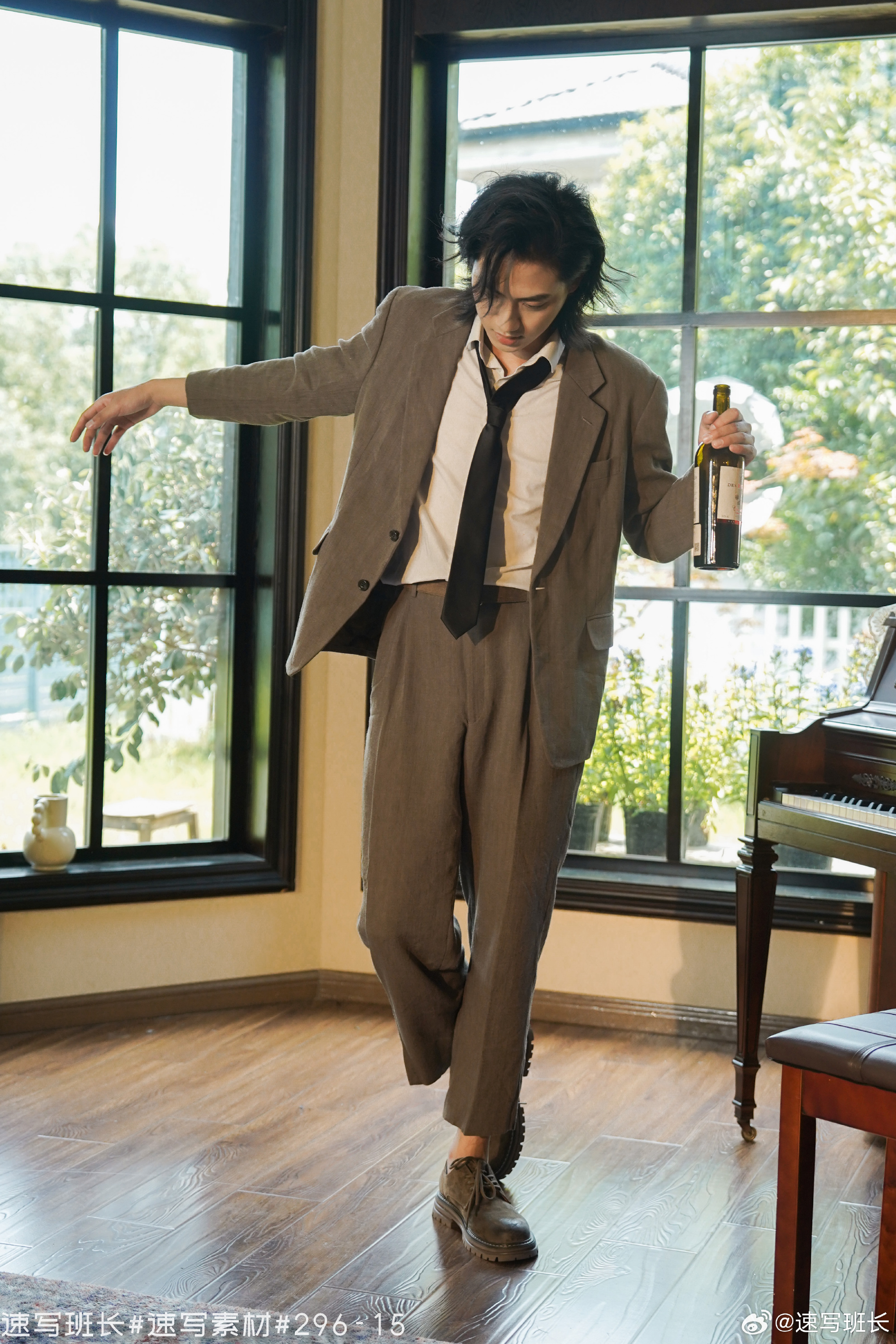 a man in a suit is dancing in a room with large windows. he is holding a wine bottle in his right hand. a piano sits in the background. the image is taken from eye level and uses natural lighting.
