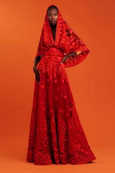 a black woman is wearing a gorgeous red dress, and she is posing for a portrait in a studio. she is wearing a red dress that is embroidered with floral motifs, the dress is elegant and flowing, and the woman's pose is graceful and confident. she is standing in front of an orange backdrop.