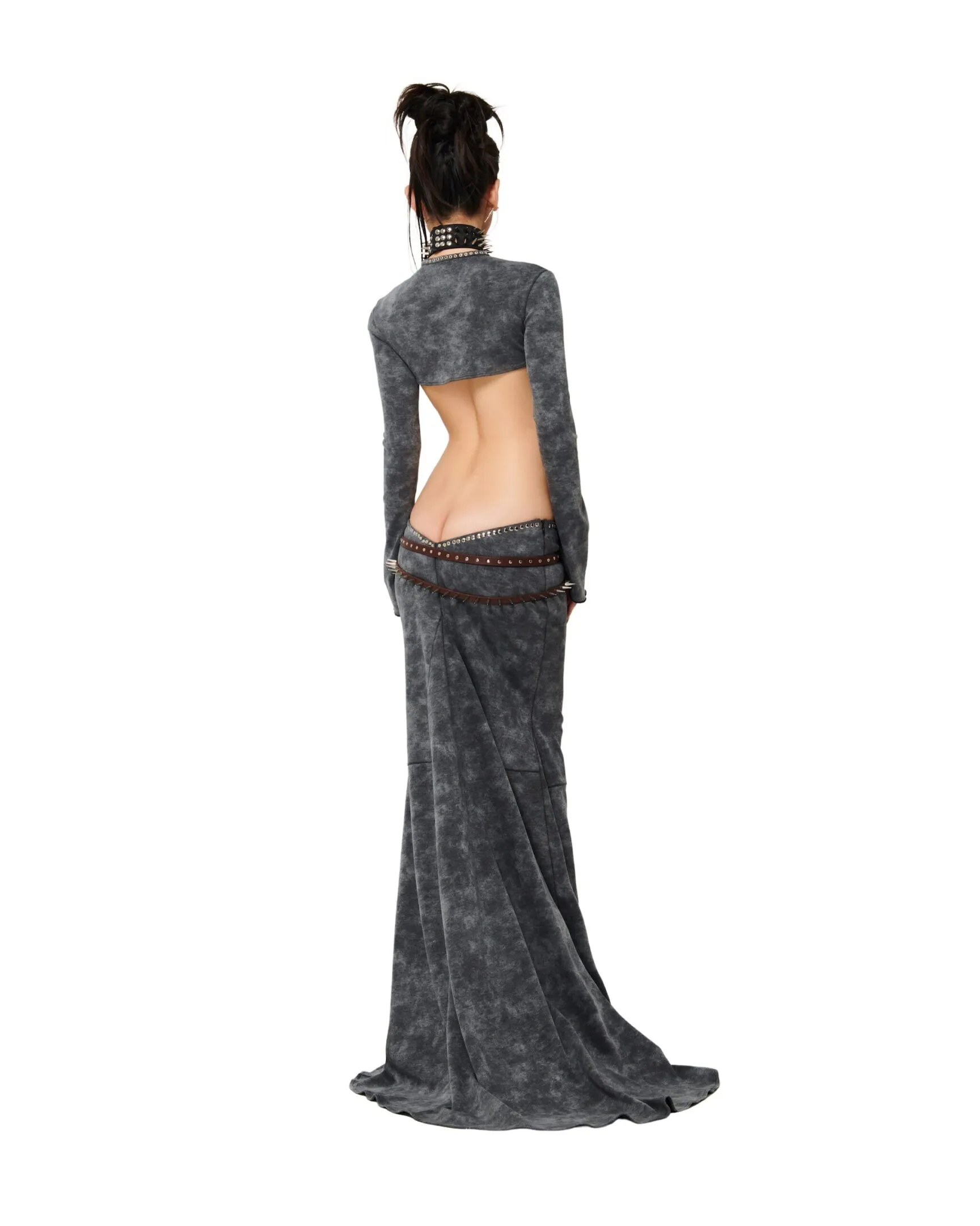 a rear view of a woman wearing a long, gray acidwash dress.  the dress features a cropped top portion that reveals a bare midriff and back. she is wearing a black choker necklace and a brown beaded belt. her hair is styled in a ponytail.  the image is taken in a studio setting against a white background.