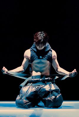a male dancer, dressed in black clothing with a ripped, shirtless torso,  is in a dynamic pose on a blue stage.  his muscles are rippling and his expression is intense.  the lighting is dramatic, casting a spotlight on the dancer, while the background is dark.  the dancer's pose and expression convey power and strength.  the water, the blue stage and the dancer's expression suggest  that this is a performance piece.  this image is a powerful statement about dedication, artistry and the power of movement.