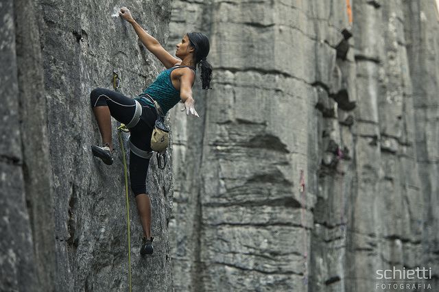 a woman is rock climbing up a steep rock face. she is wearing climbing gear and is using her hands and feet to grip the holds. the rock face is gray and there are some trees in the background.