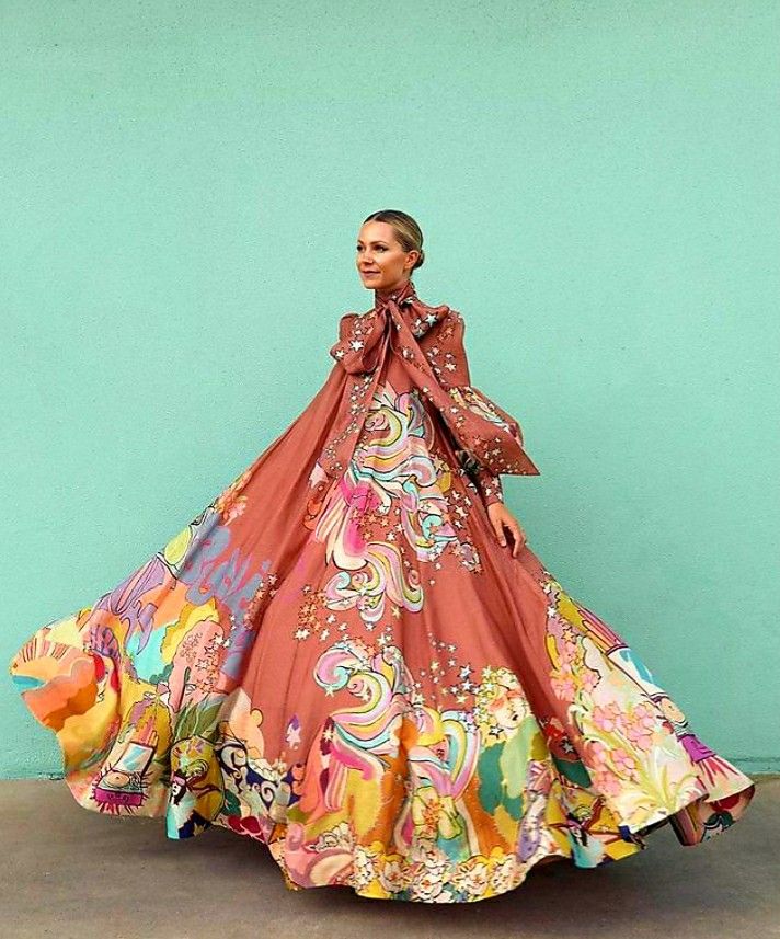 a woman is standing in front of a light blue wall wearing a large, colorful dress with a bow at the neck. the dress features an elaborate print with various colors and patterns. she is posing for the photo, and the image captures the full length of her outfit.