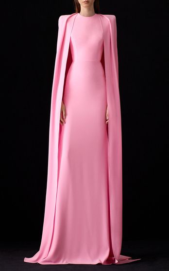 the image shows a woman wearing a stunning pink maxi dress with a cape. the dress is floorlength, longsleeved, and has a fitted silhouette. the cape is attached to the shoulders and flows elegantly down the back. the model stands against a plain black backdrop, highlighting the dress's vibrant color and exquisite design. the overall look exudes elegance and sophistication, suitable for a formal occasion like a gala or redcarpet event.