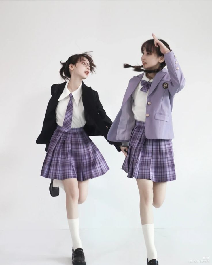 two young girls in school uniform are standing and posing for a picture. they are wearing plaid skirts, blazers, white shirts, and ties. they are both smiling and look cute.