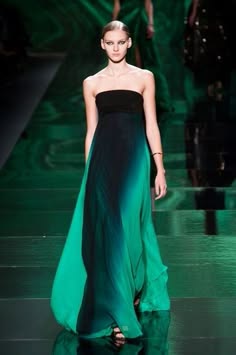 a female model walks down a runway in a long, flowing, ombre dress. the dress is a deep green color at the bottom, fading to black at the top. the model has her hair pulled back and is wearing minimal makeup. the background is a green and blue backdrop.