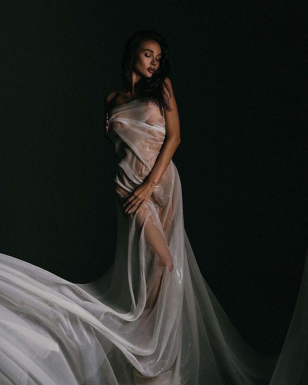 the image is a stylized portrait of a woman.  she is posed gracefully, her body mostly veiled by a sheer, flowing white fabric that drapes around her like a gown. the fabric is translucent, hinting at her form underneath, creating a sense of both intimacy and mystery. her dark hair contrasts with the light fabric, and her expression is serene and somewhat introspective. the background is dark and plain, drawing all attention to the model and her pose.  the overall mood is sensual, elegant, and artistic. the wet look of the fabric suggests a possible water theme. the lighting is dramatic, using shadows and highlights to accentuate the woman's form and the fabric's texture.