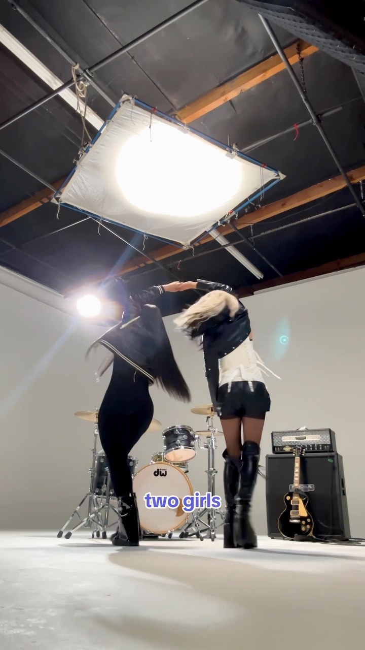 the image shows two girls dancing in a music studio, with a drum set, guitar, and amplifier visible in the background. there's a large overhead light illuminating the scene.