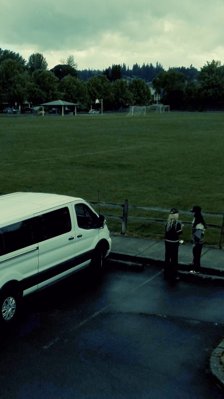a medium shot of a white van parked in a parking lot, next to a grassy field. two people are standing near the van, seemingly having a conversation. in the background, there are trees, a soccer goal, and an overcast sky.