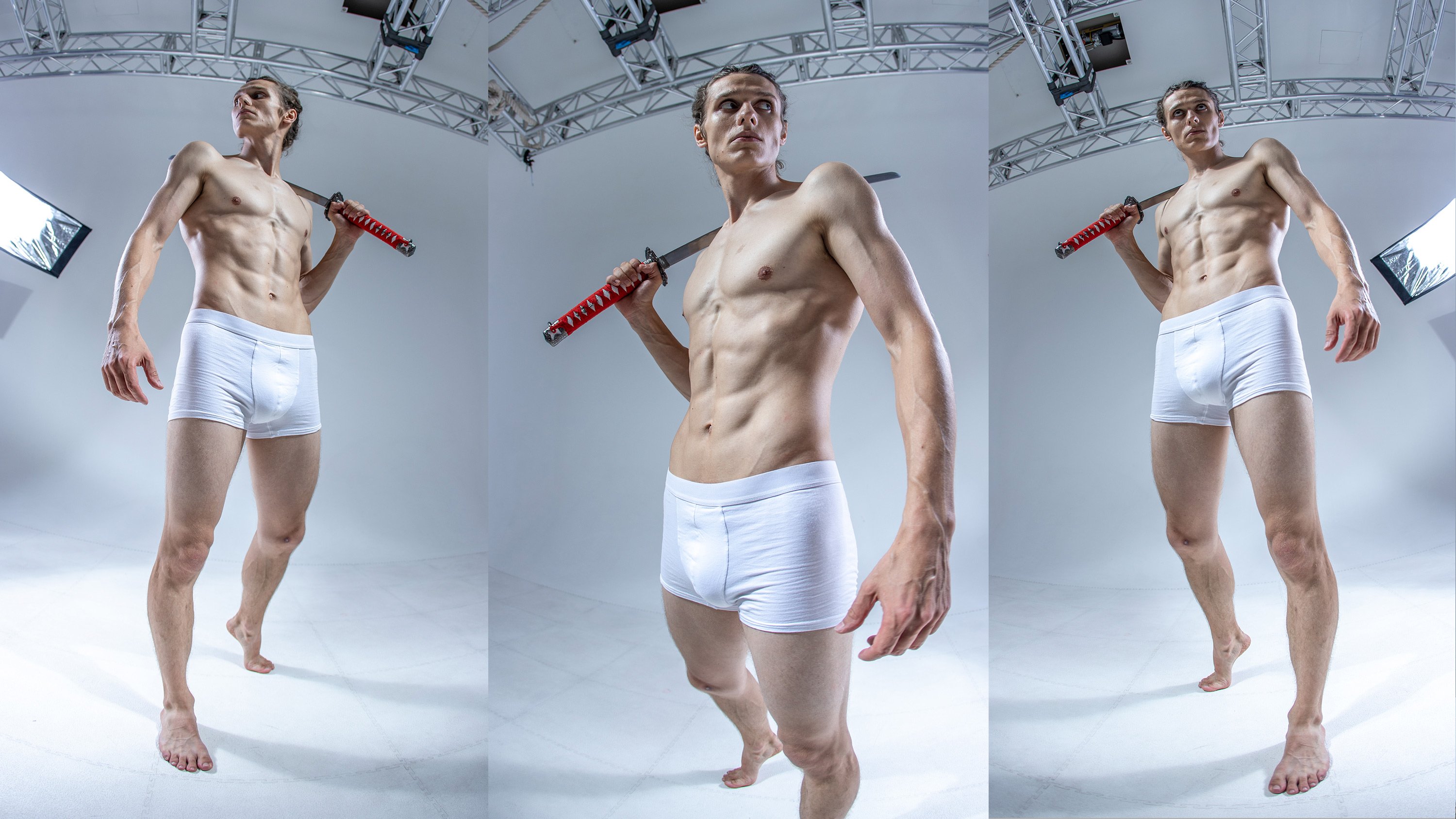 a muscular young man stands in a studio, holding a katana sword, looking to his right. he is wearing white boxer briefs and is positioned with his left leg bent and his right leg straight. the photo appears to be taken using a fisheye lens.