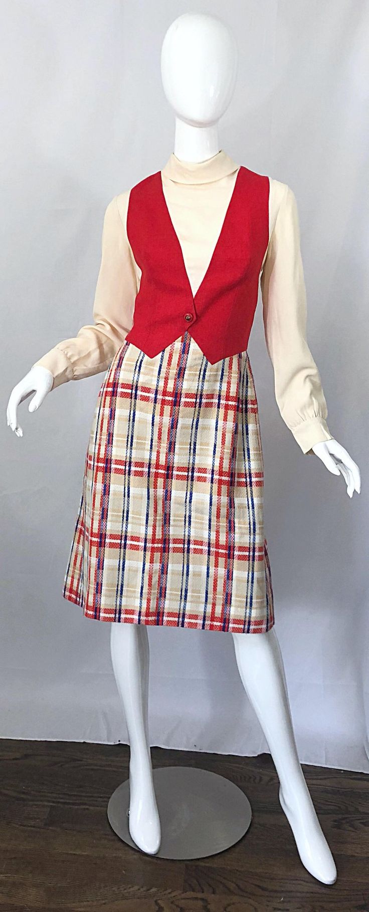 a white mannequin is dressed in a 1970s outfit. it includes a cream turtleneck, a red vest, and a plaid skirt. the background is plain white and the lighting suggests that the photo was taken in a studio.