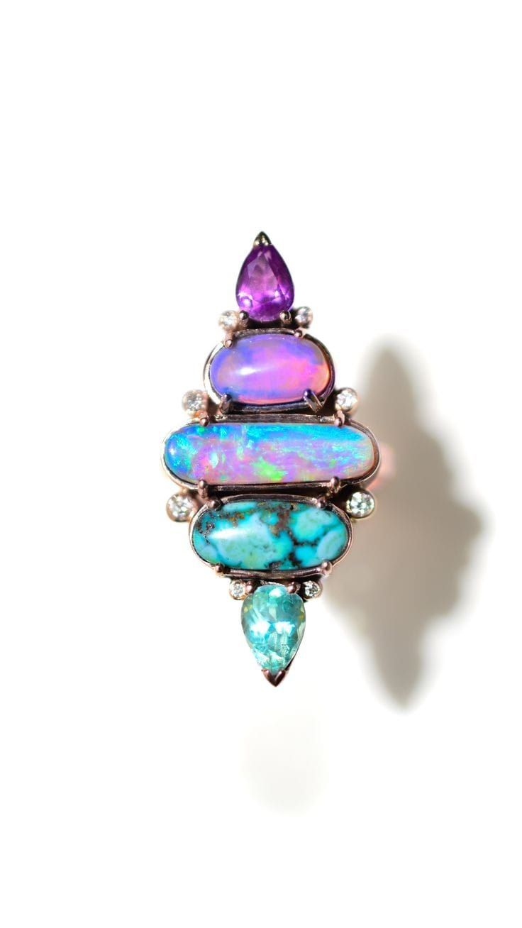 a closeup image of a beautiful ring with a variety of gemstones, including a pink opal, a turquoise stone, and a blue aquamarine.