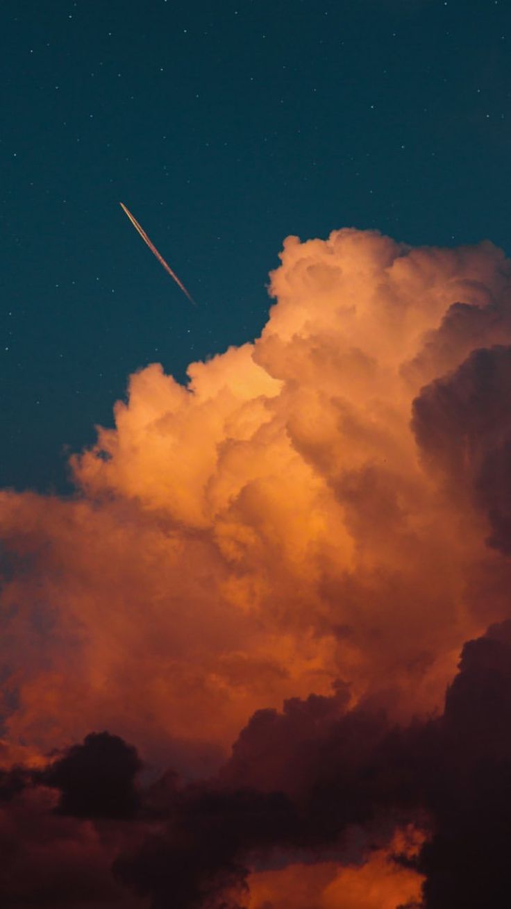 the image shows a vibrant sunset scene with large, orangetinted clouds taking up most of the frame. at the top of the image, a dark blue sky filled with stars is visible. an airplane trail streaks across the dark sky.