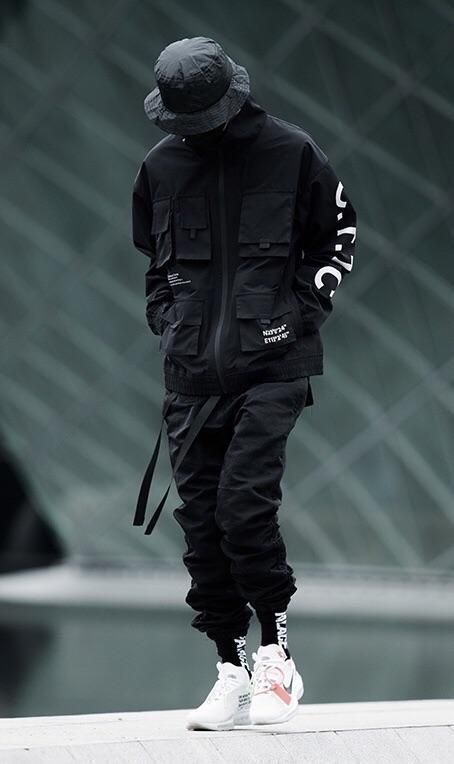 a man is wearing a black jacket with a white print, black pants, a black hat and white sneakers. he's standing in an urban setting.