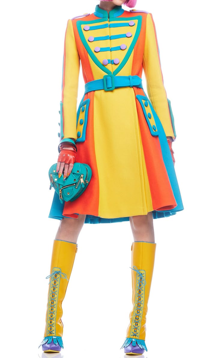 a model is standing on a white background, wearing a colorful coat with yellow, orange, and blue panels, a matching belt, and yellow kneehigh boots with blue accents. she is holding a blue heartshaped handbag and wearing red gloves.