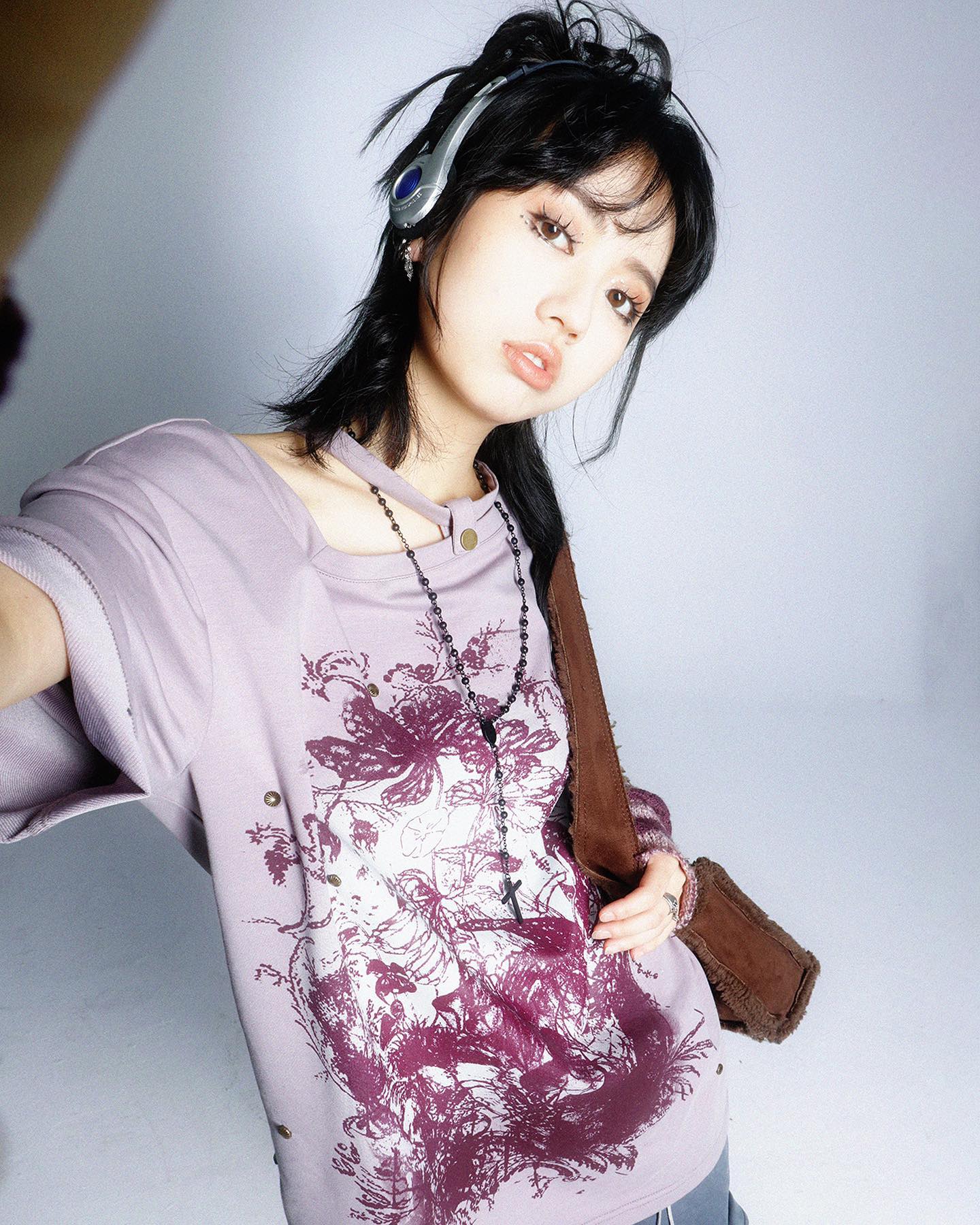 loseup selfiestyle portrait of a young asian woman with dark layered hair and bangs. she's wearing a loosefitting, lavendercolored tshirt with a dark, vintagestyle floral and possibly creature graphic print. the shirt has distressed detailing. she also has on a dark beaded necklace resembling a rosary, and a brown crossbody bag. the woman is wearing silver overear headphones. the background is plain white, creating a stark contrast to the woman's attire and giving a focus to her expression and clothing details. the overall style is casual, with grunge and vintage influences.