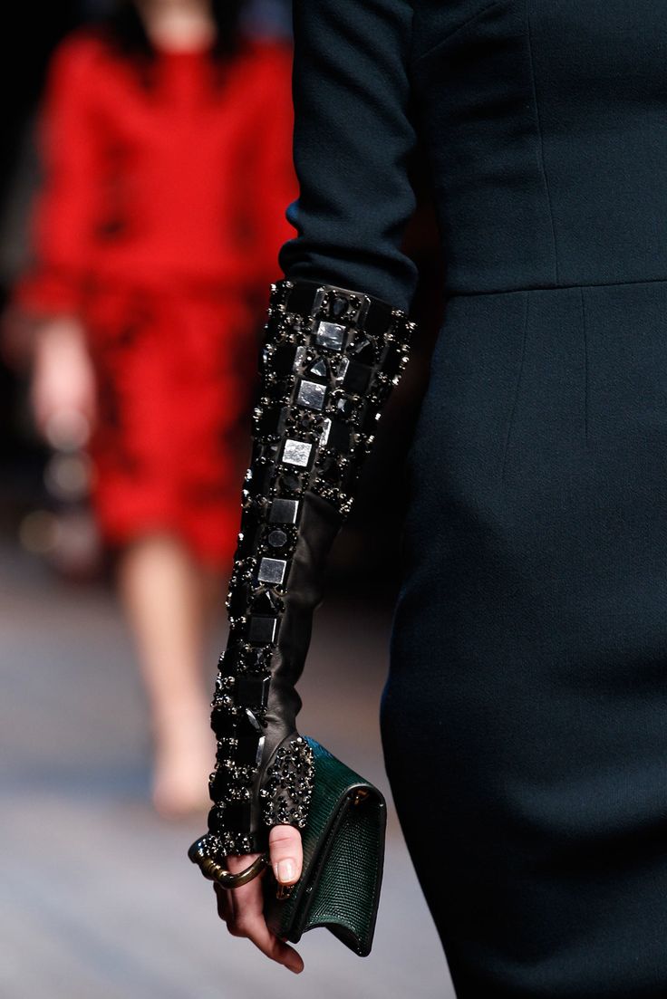 loseup shot of a model's arm and hand showcasing a long black glove adorned with decorative black and silver embellishments. the model is holding a dark green handbag. the background is blurred, showing another model in a red dress. the focus is on the intricate details of the glove and handbag, highlighting the luxurious and stylish elements of the outfit. the overall ambiance suggests a highfashion runway setting.