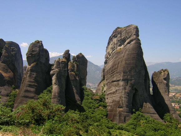 the image showcases the unique rock formations of meteora, greece. towering sandstone pillars dominate the landscape, with lush greenery covering the ground and mountains visible in the background. the sky is a clear blue, adding to the scenic beauty of the area.